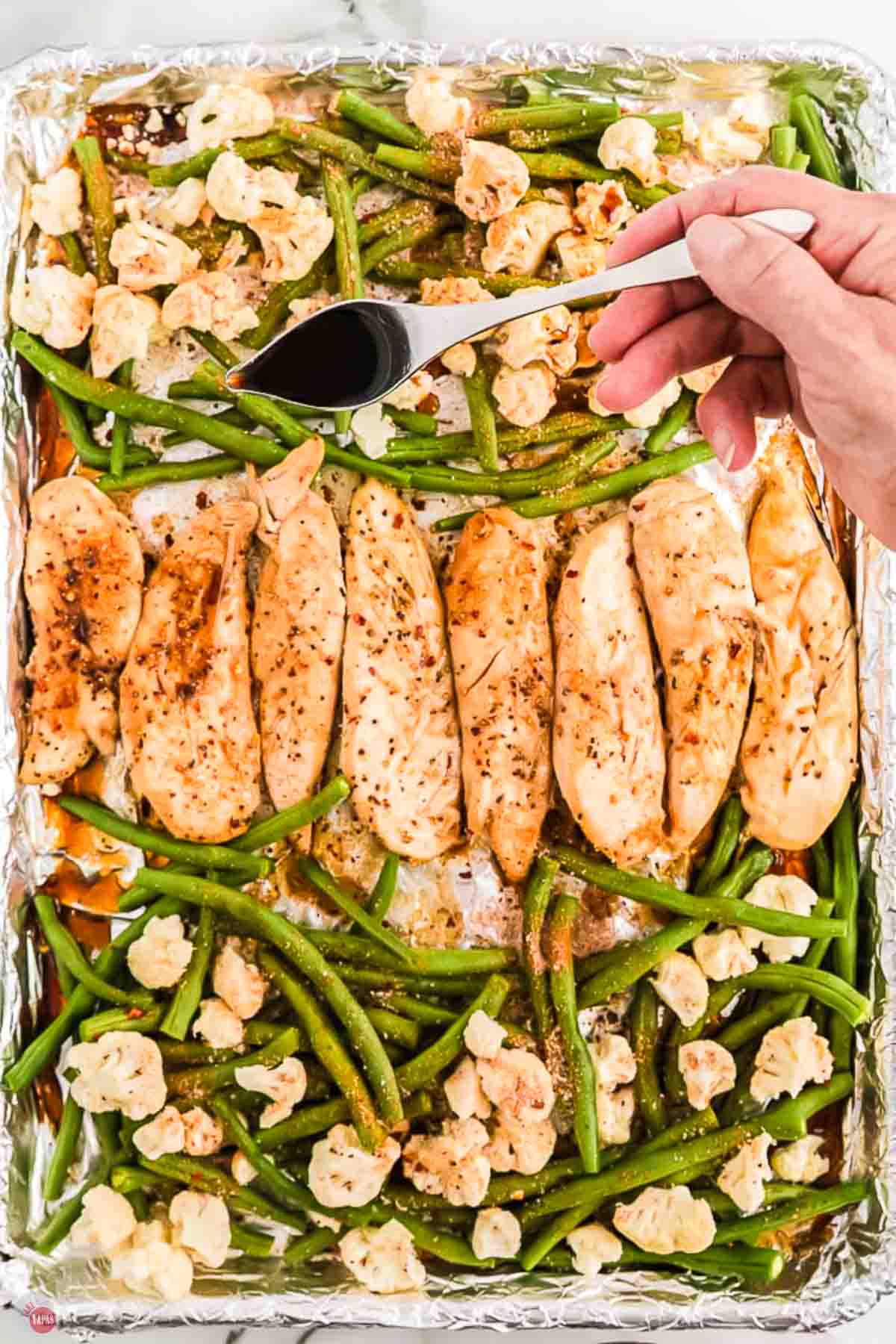 baked chicken and vegetables on a baking sheet with a spoon drizzling sauce on top
