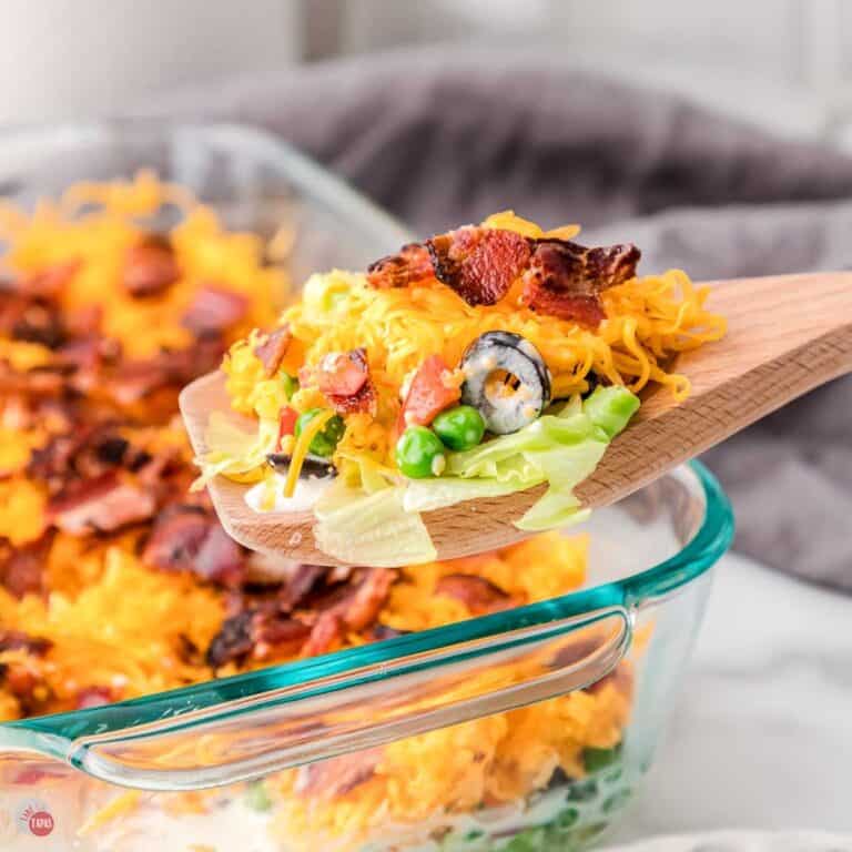 layered salad in a clear dish with a wood spoon scooping some out