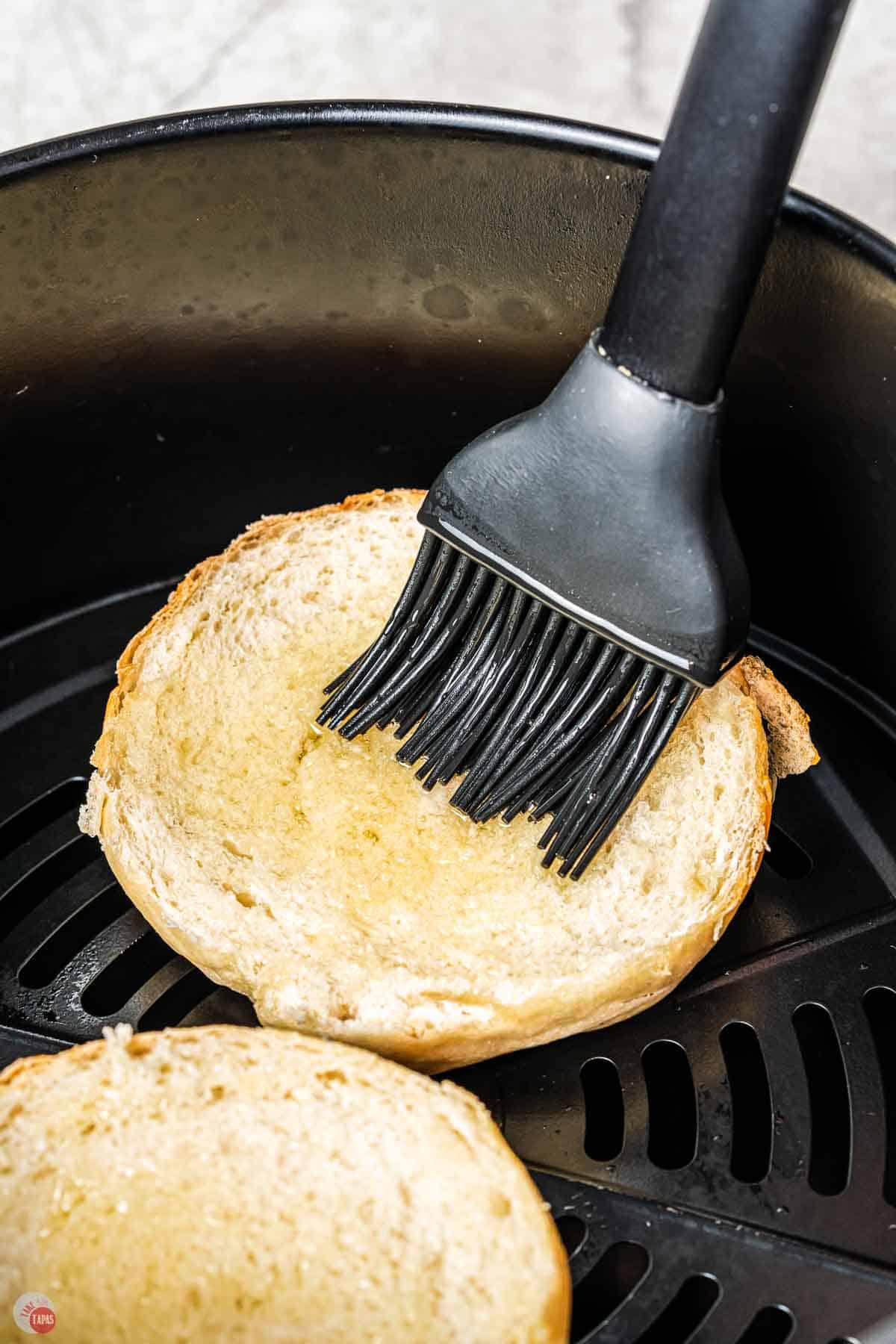 sliced bagel in an air fryer basket with a silicone brush applying olive oil