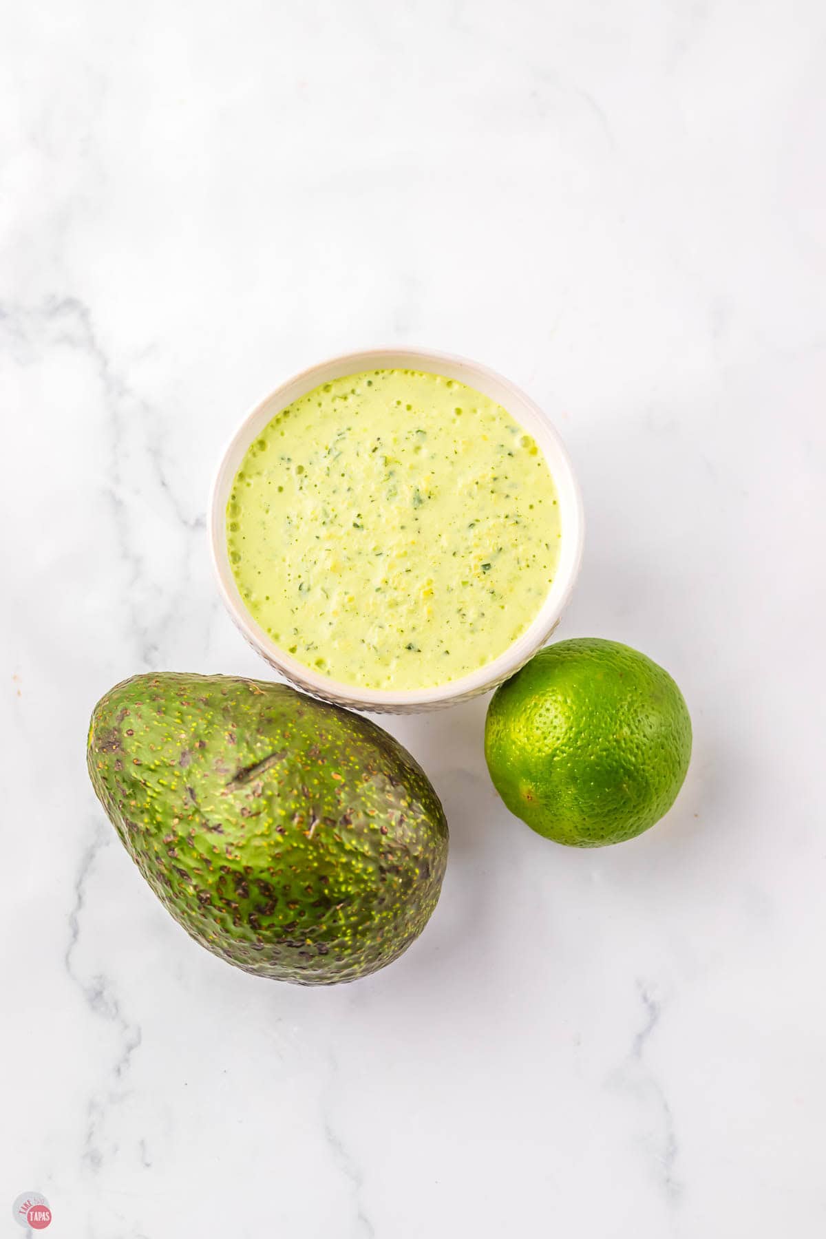 white bowl of avocado ranch dressing next to an avocado and a lime