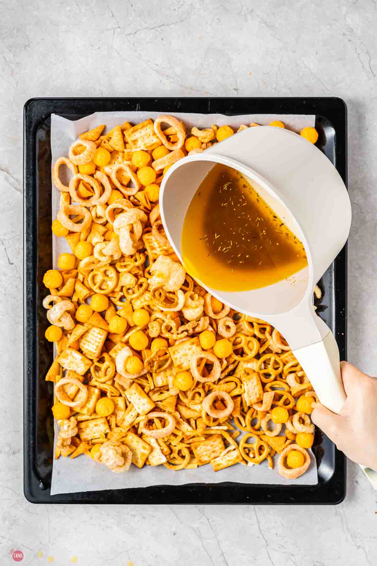 sauce being poured over chex mix on a baking sheet