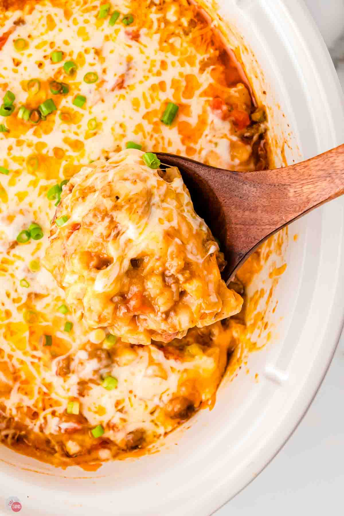 close up of taco casserole in a white slow cooker being scooped out with a wood spoon
