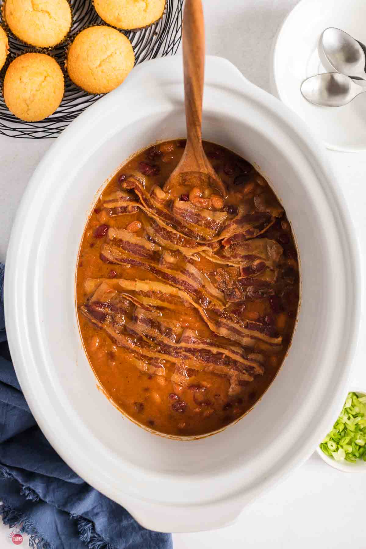 slow cooker with beans, bacon, and a wood spoon