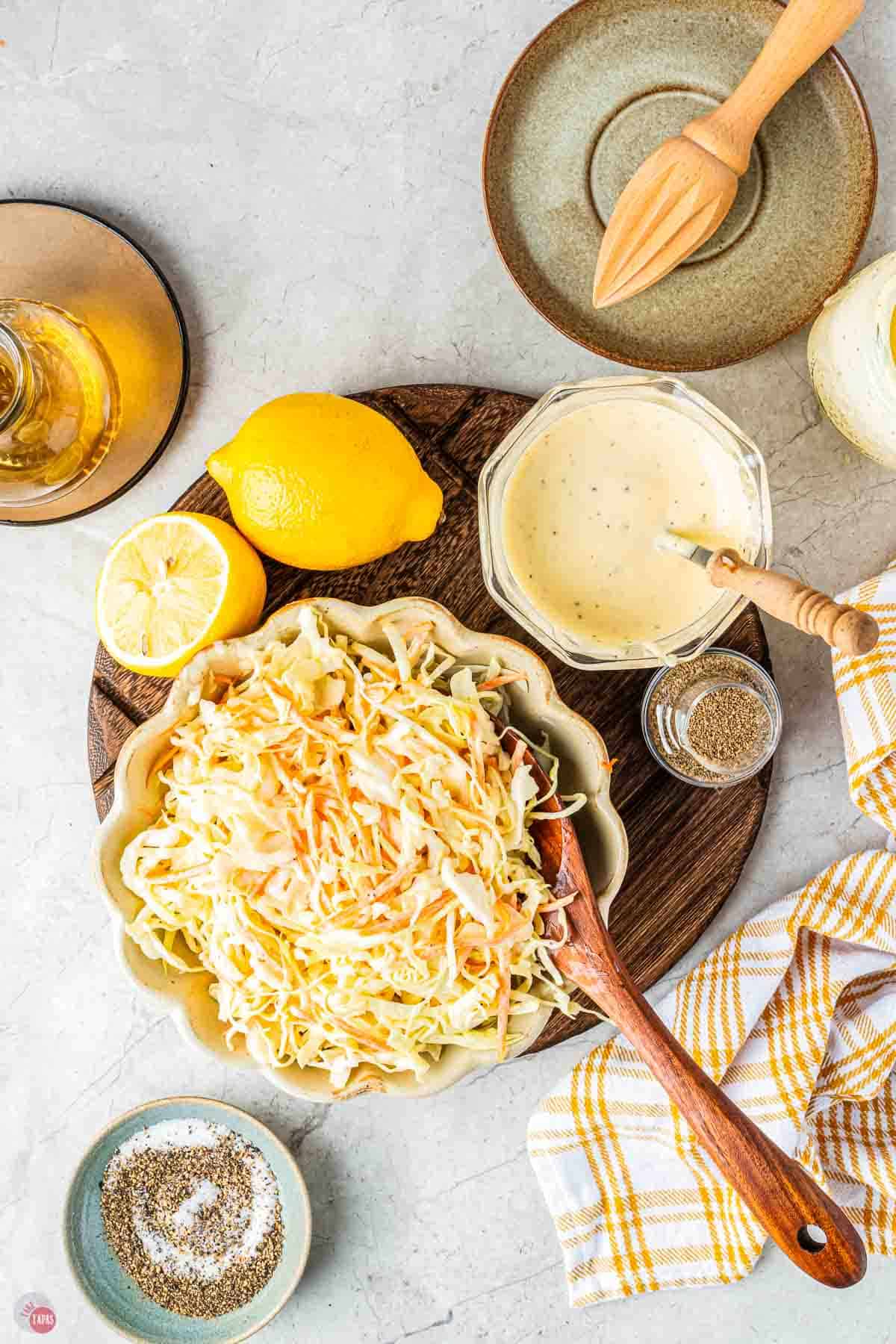 wood board with bowl of coleslaw, jar of dressing, and a few lemons