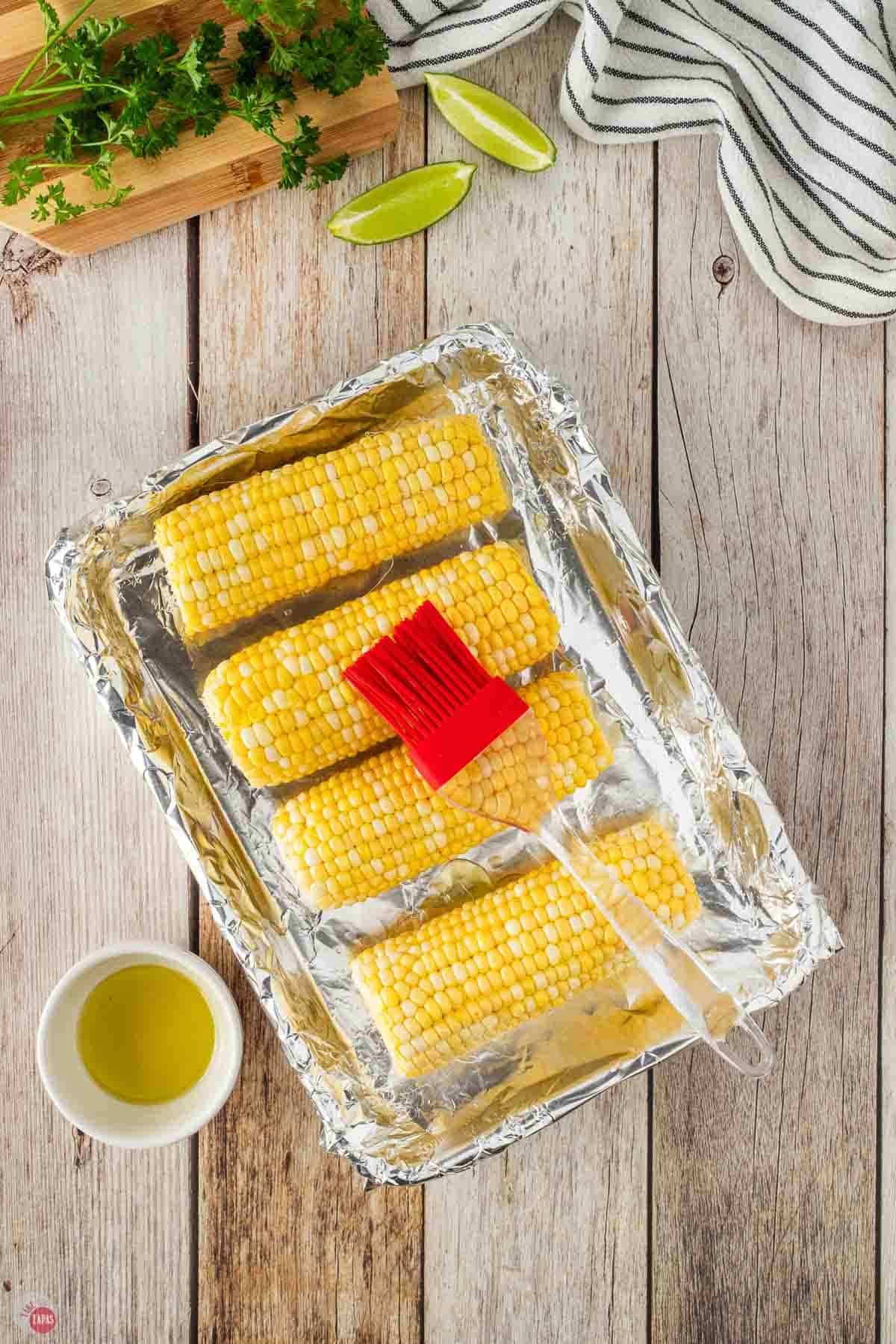 4 cobs of corn with a red silicon brush brushing on olive oil