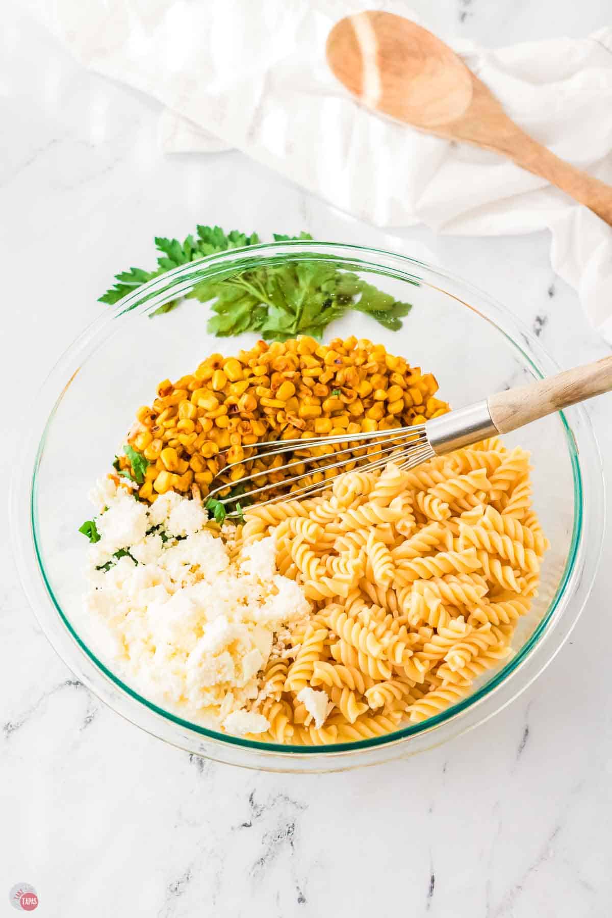 clear bowl with rotini pasta, roasted corn, and cheese with a wood spoon