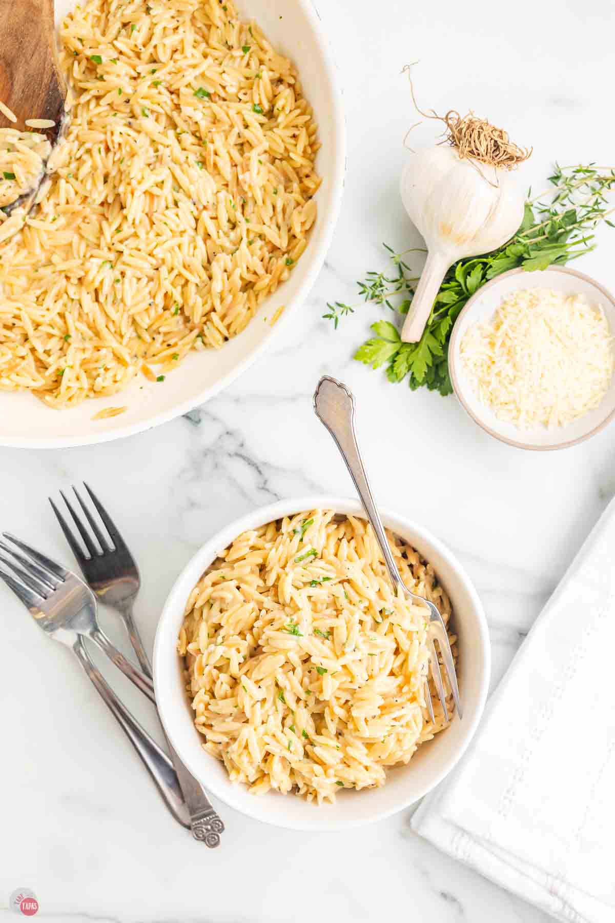 two bowls of pasta with a bowl of shredded cheese and forks next to it
