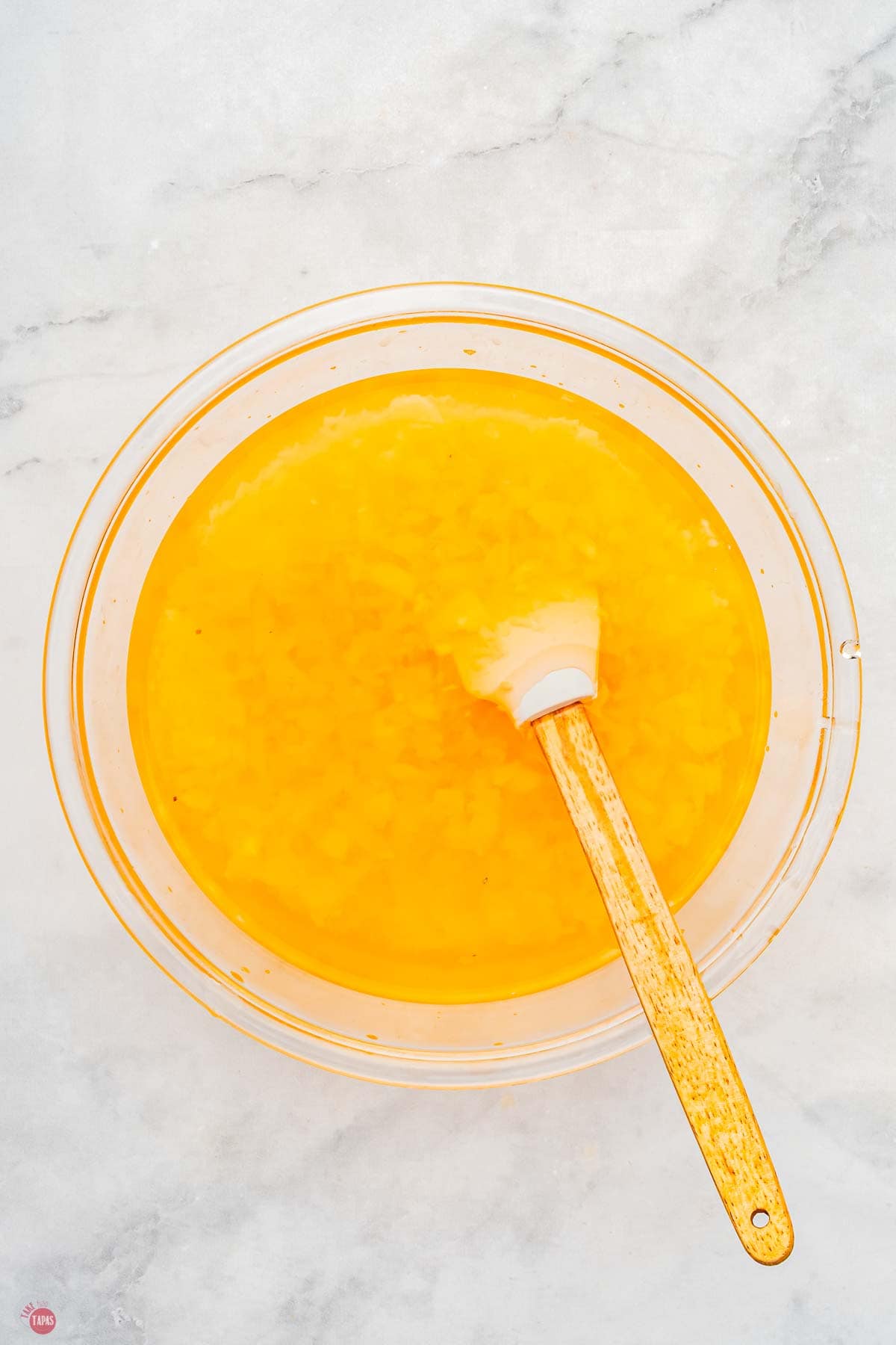 pineapple jello being stirred in a clear bowl with a spatula