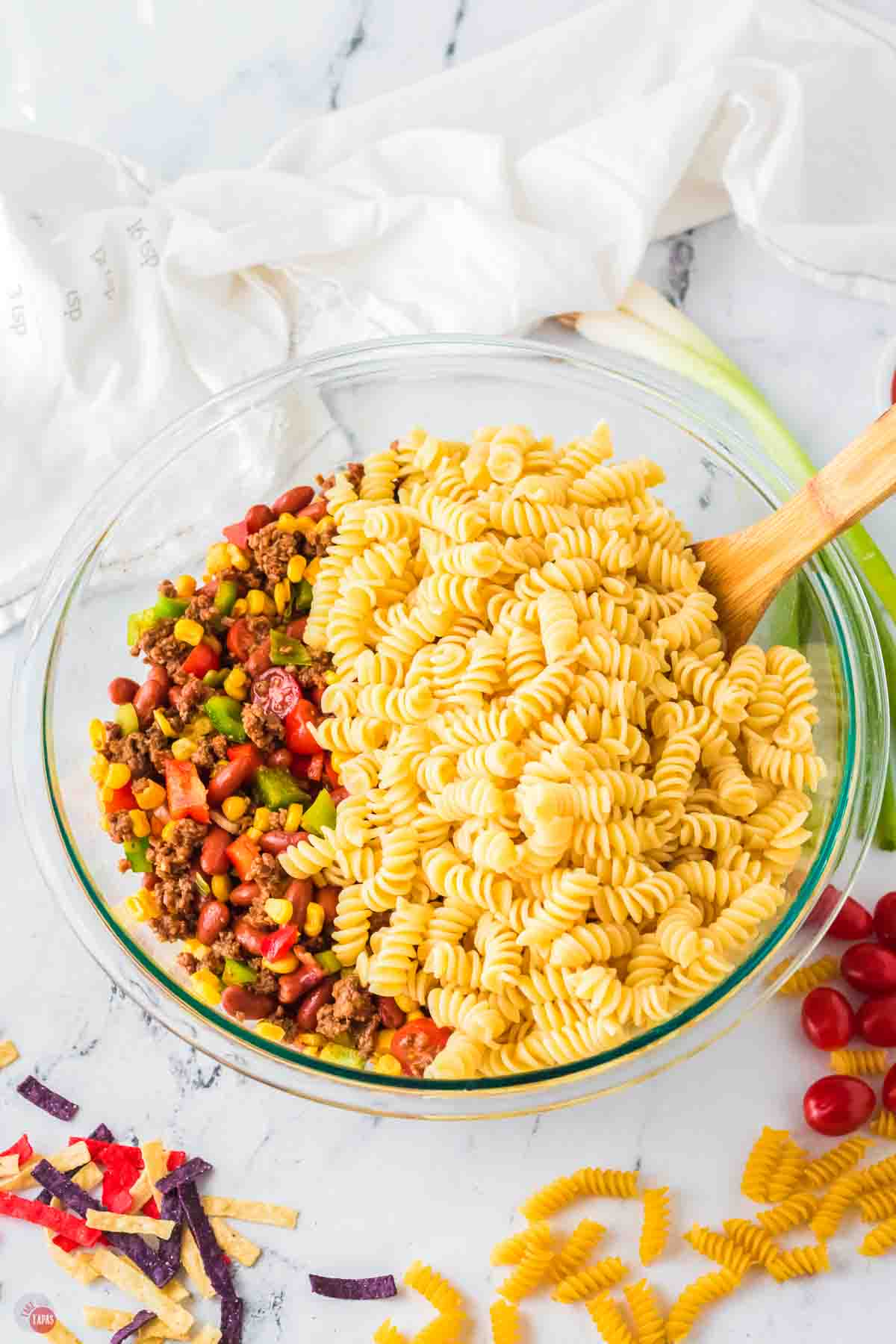 clear bowl with chopped vegetables and cooked rotini pasta being stirred with a wood spoon