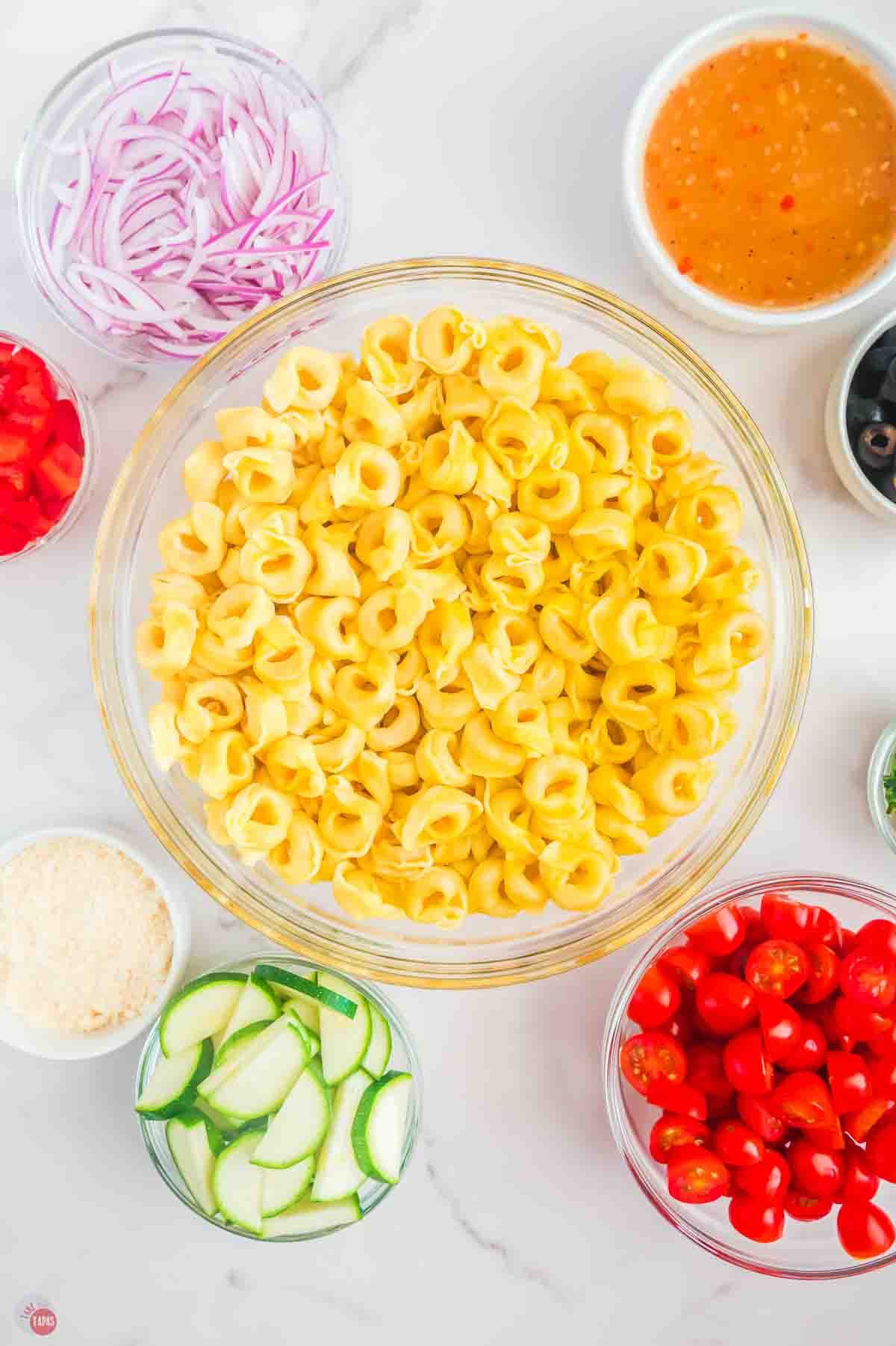 cooked cheese tortellini in a large clear bowl surrounded by smaller bowls of chopped vegetables