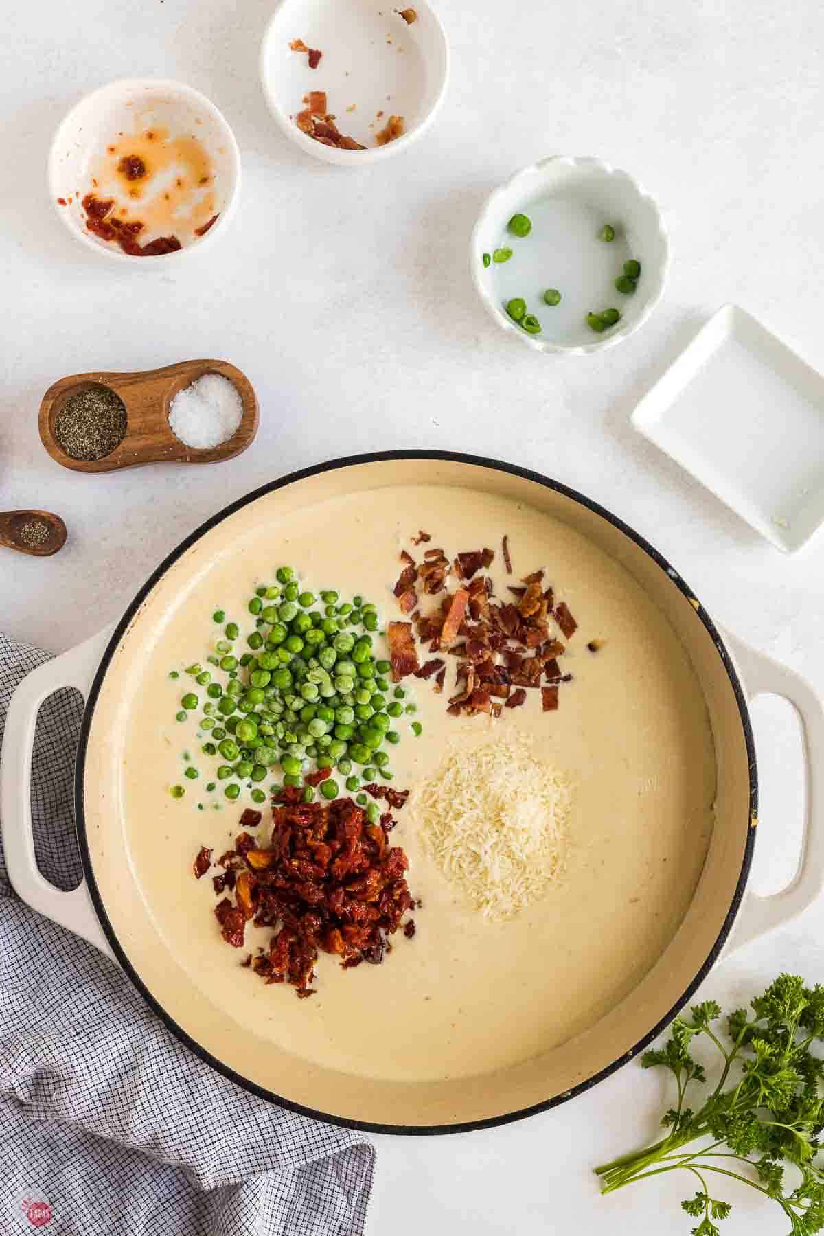 white skillet with heavy cream, bacon, peas, and sun-dried tomatoes
