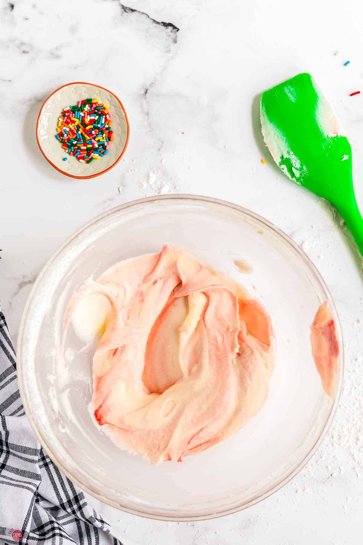pink frosting mixing in a clear bowl with a green spatula