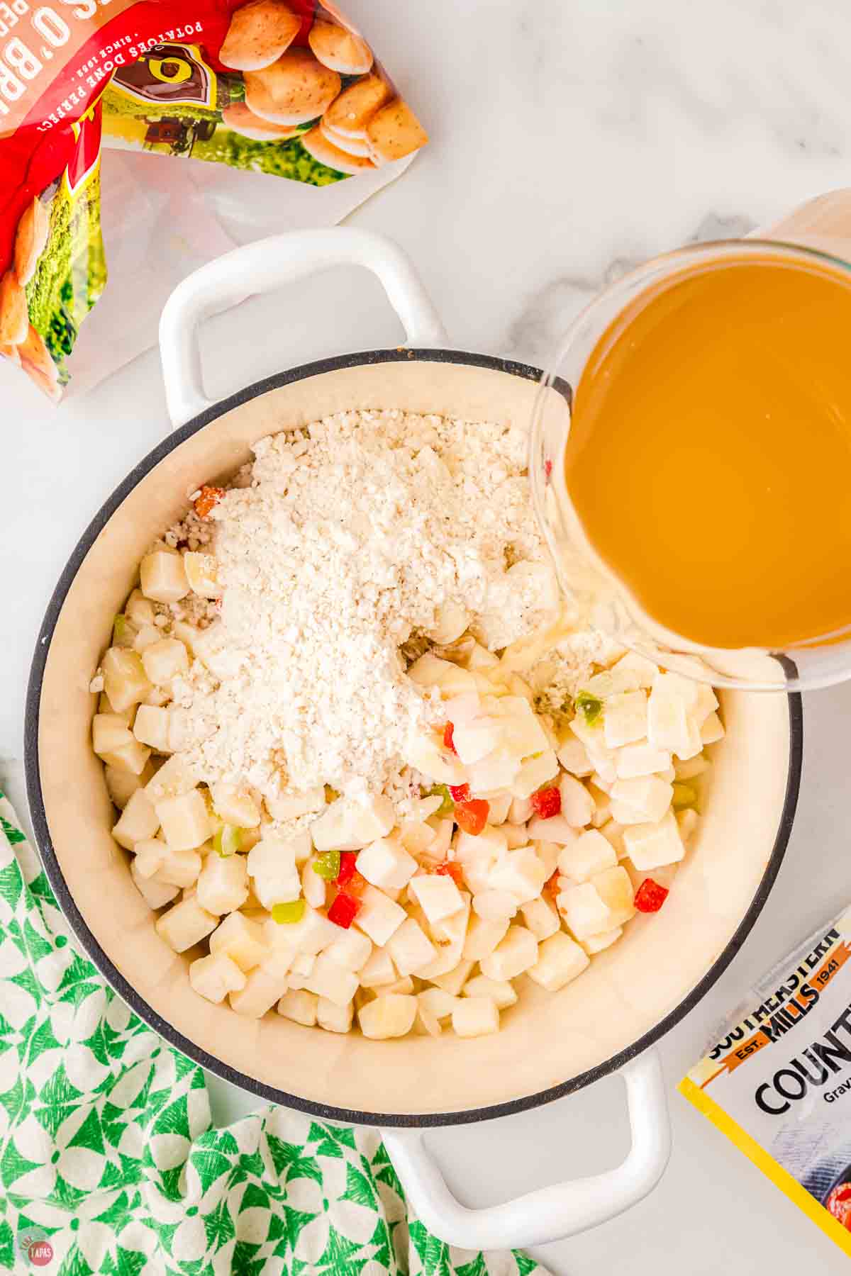 white soup pot filled with frozen diced potatoes and gravy mix with broth being poured in
