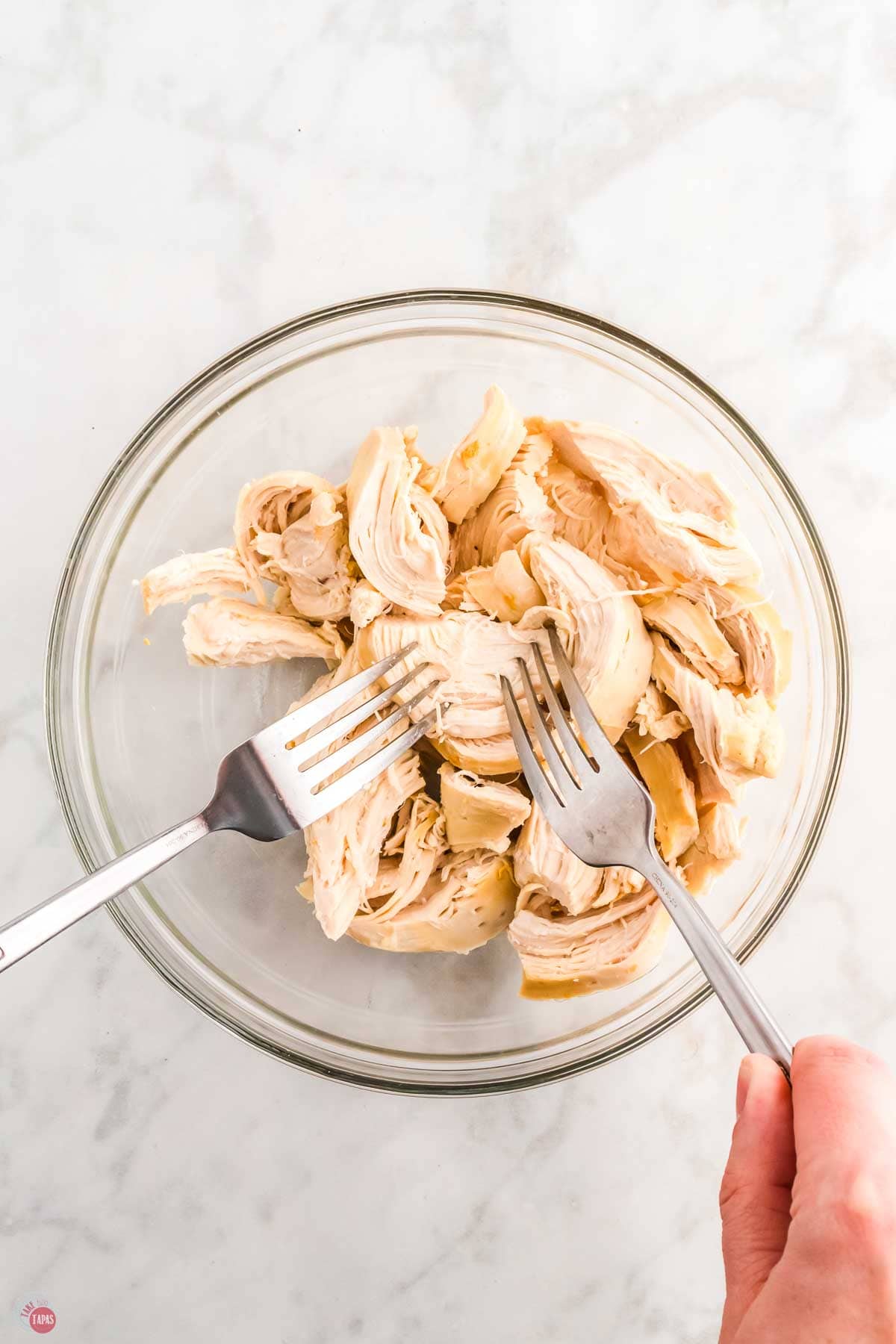 chicken meat being shredded with forks in a clear mixing bowl