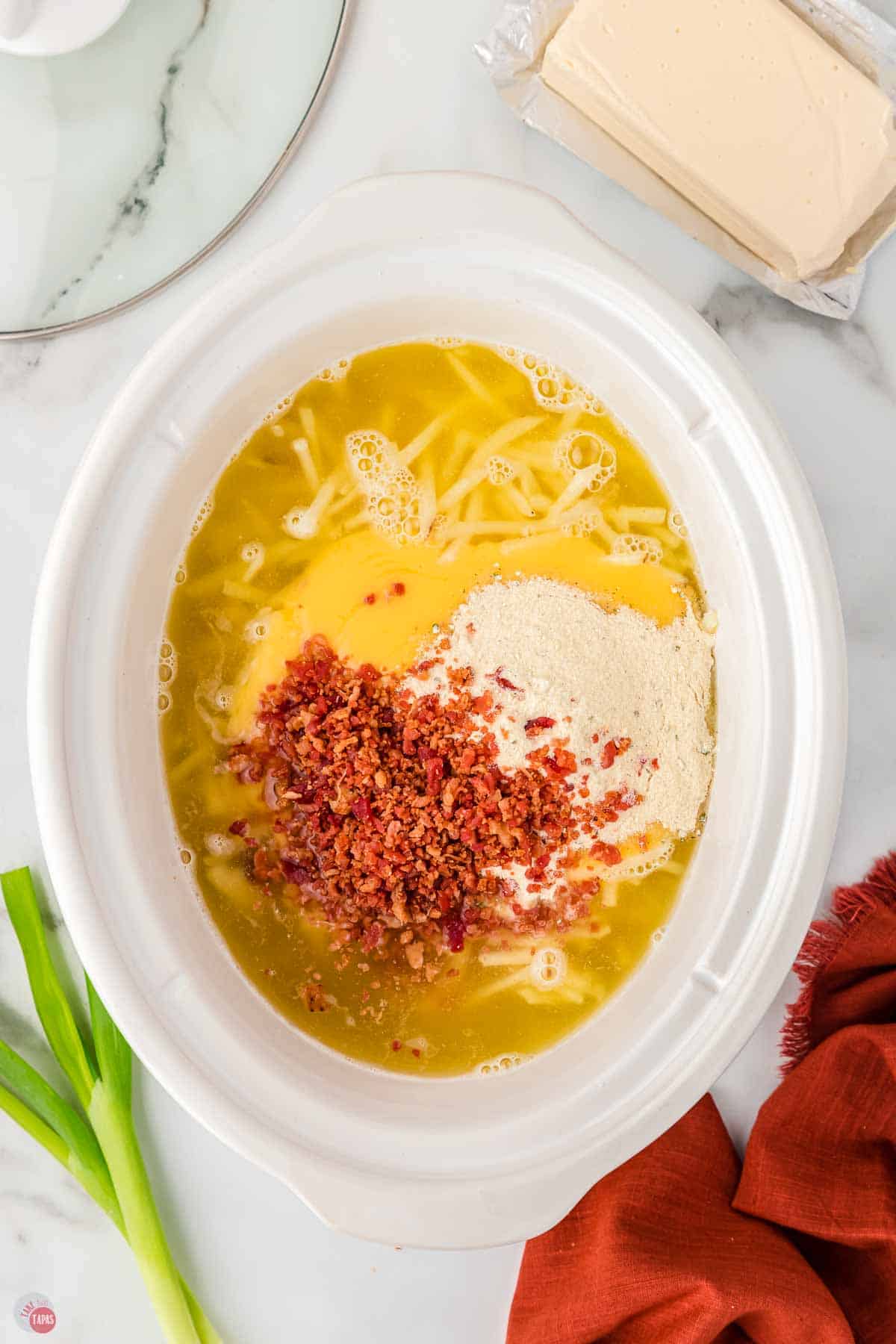 white slow cooker bowl filled with hashbrowns, broth, bacon, and seasoning