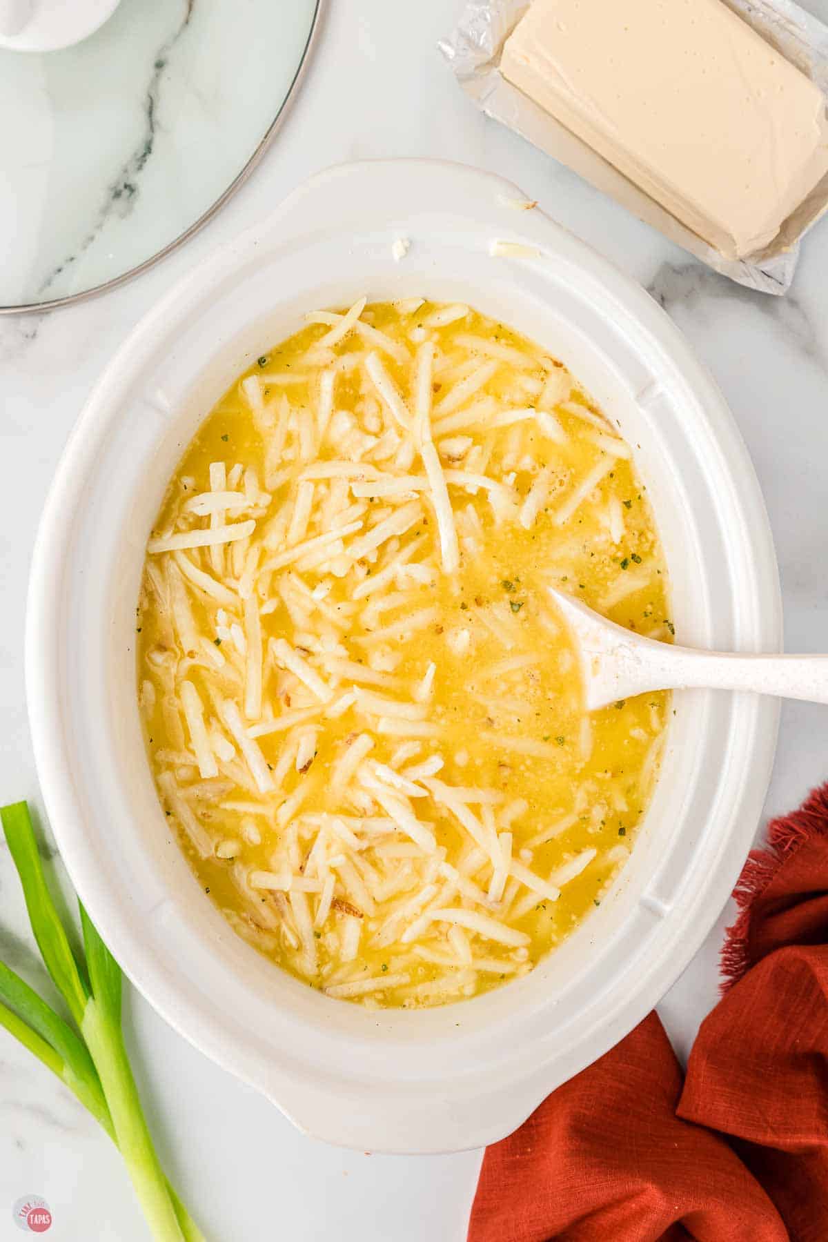 slow cooker full of hashbrowns and broth being stirred with a white spoon