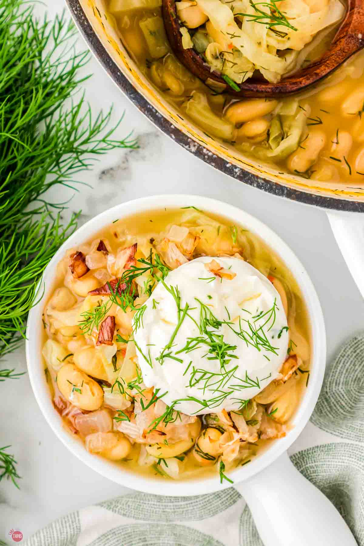 bowl of bean soup topped with dollop of sour cream next to a soup pot and a bunch of fresh dill