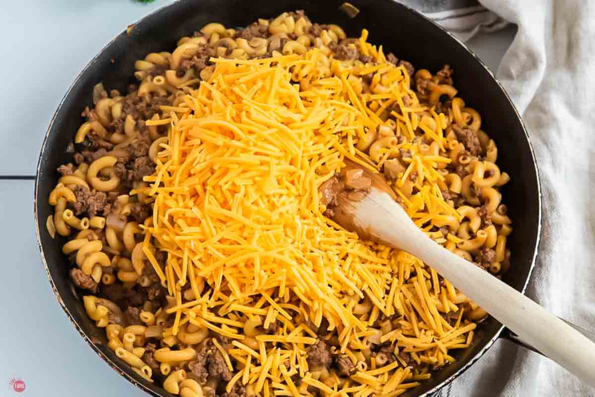 skillet with macaroni and ground beef with shredded cheese being stirred in