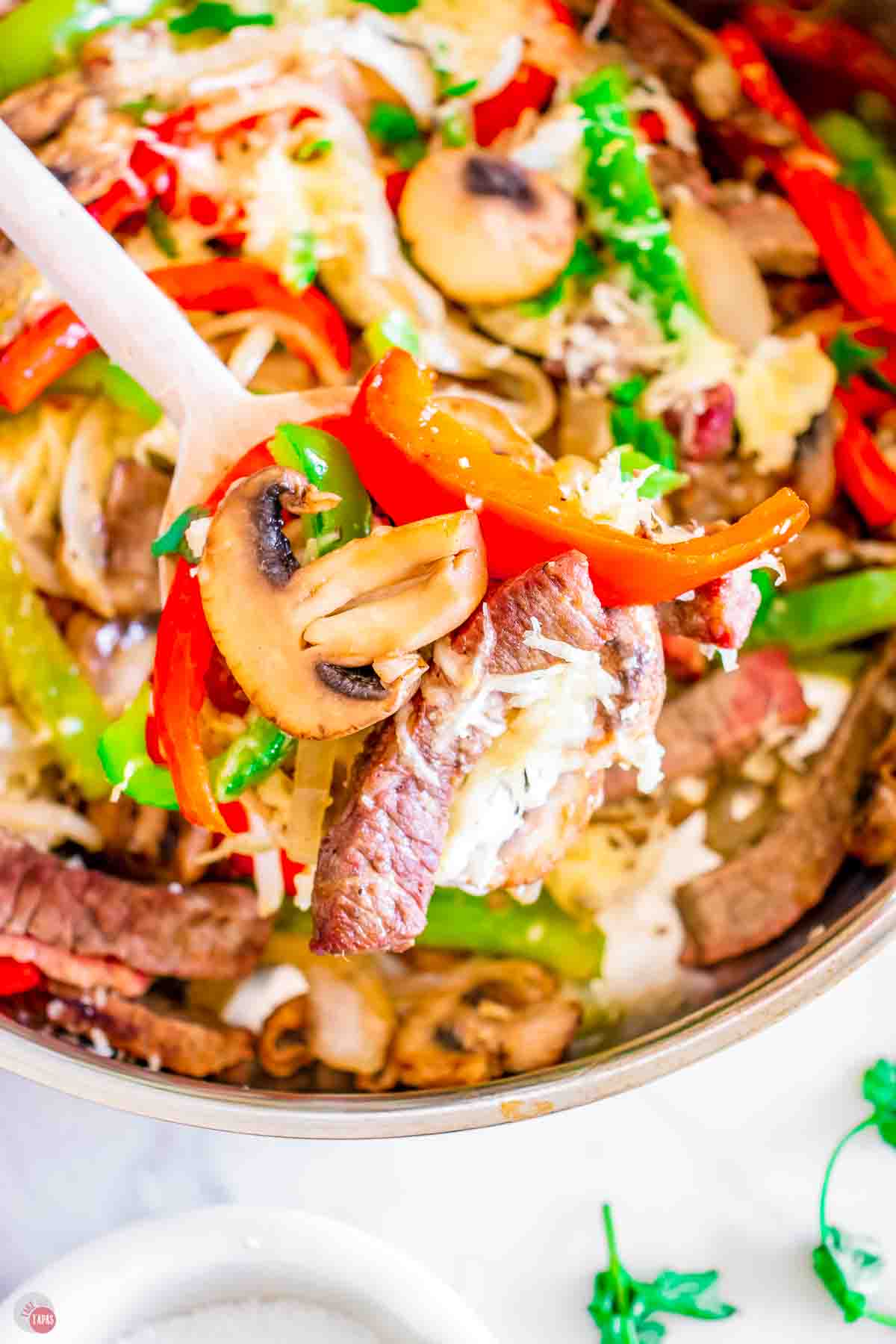 close up of skillet with a white spoon scooping mushrooms, peppers, and onions