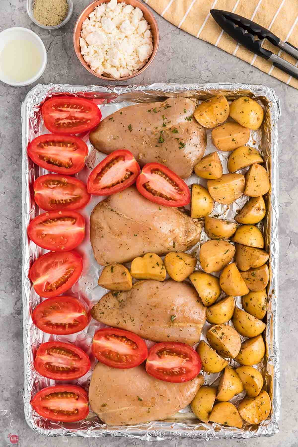 raw chicken breasts, potatoes, and halved tomatoes on a baking sheet lined with aluminum foil