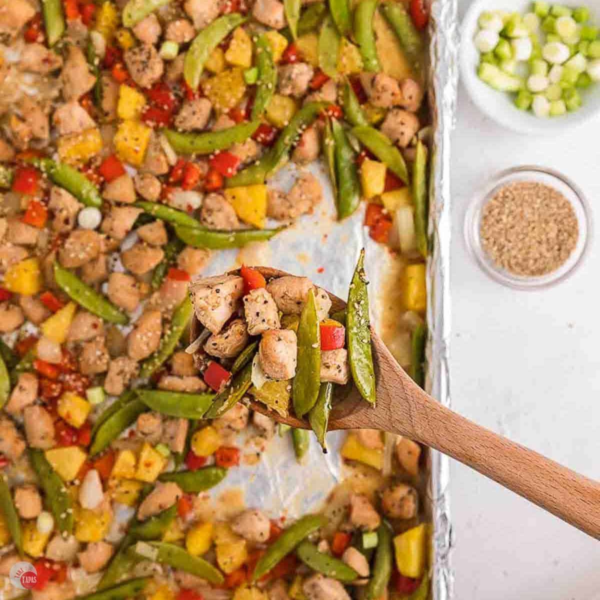 close up of chicken and vegetables in a wood spoon hovering over a sheet pan