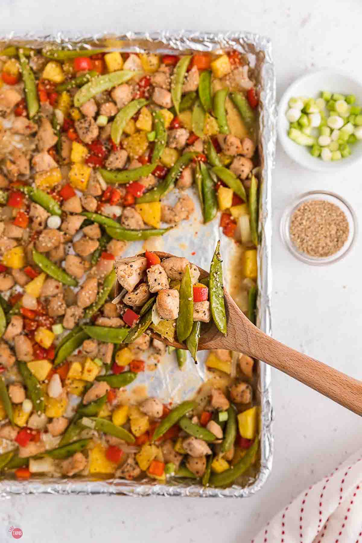 close up of chicken and vegetables in a wood spoon hovering over a sheet pan
