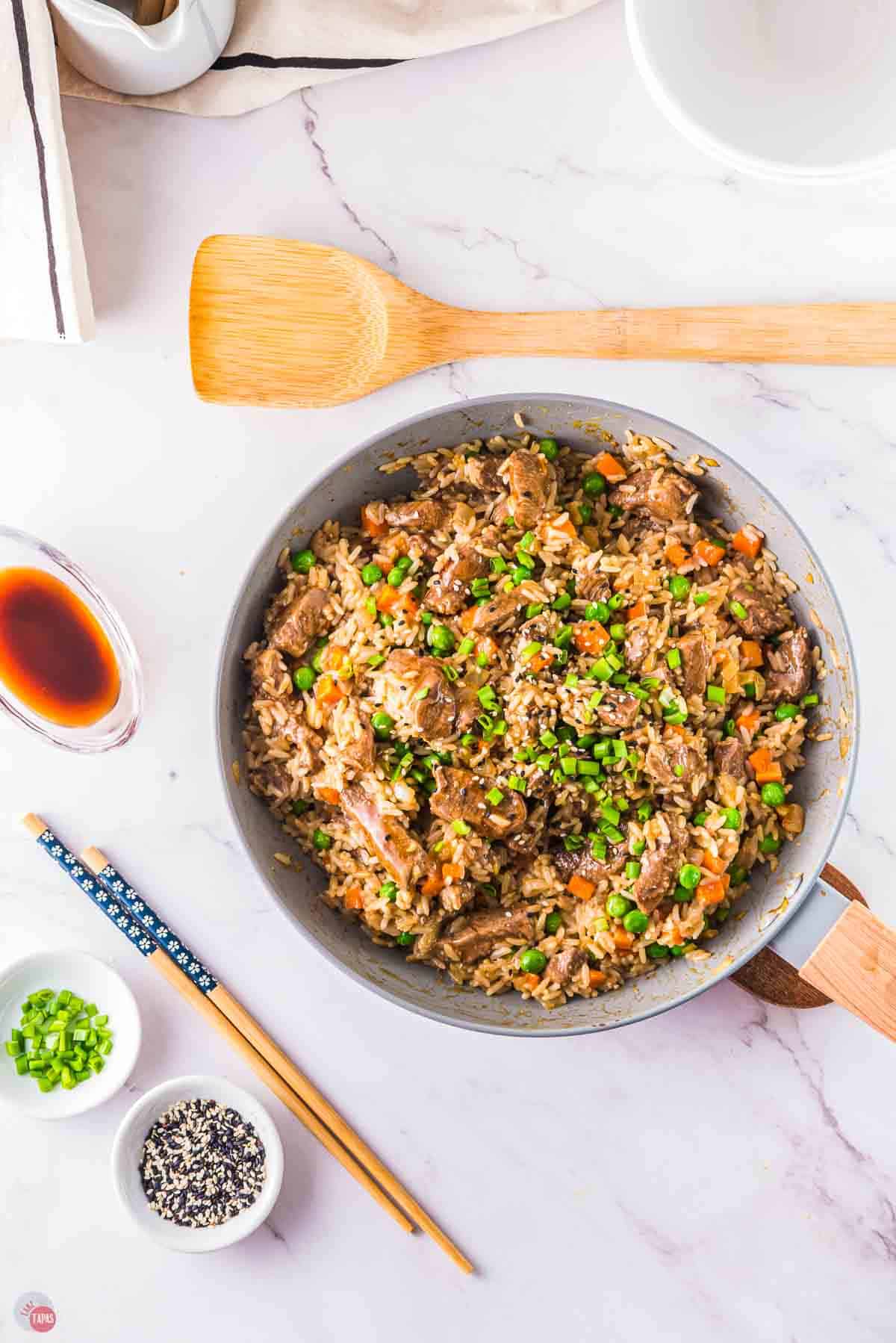 overhead picture of skillet with rice and meat in it next to a wood spoon and a small bowl of soy sauce