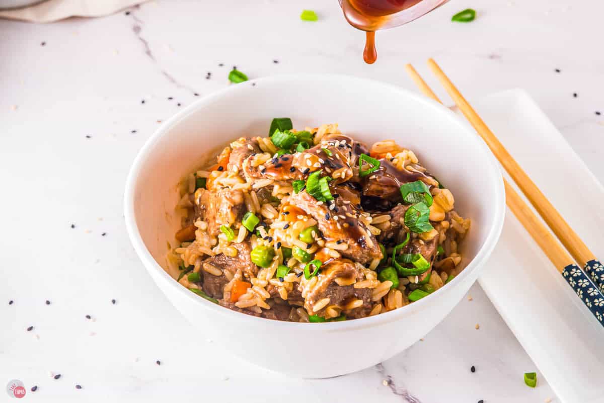 white bowl of fried rice with steak and  soy sauce being poured over the top of it