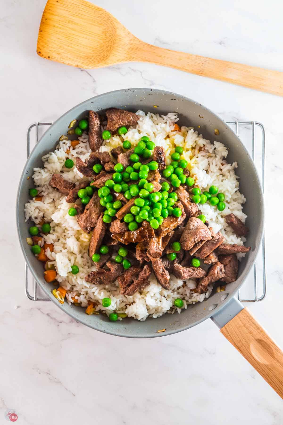 white rice in a skillet topped with cooked strips of steak and green peas