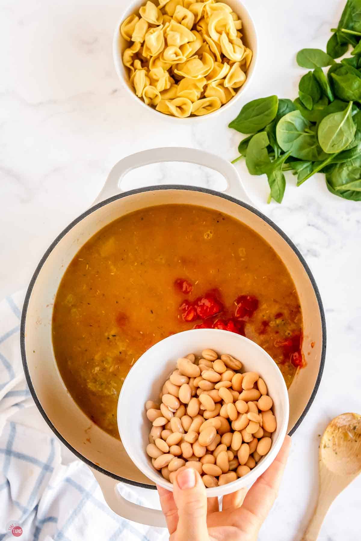 soup pot of simmering broth with a bowl of white beans being poured in