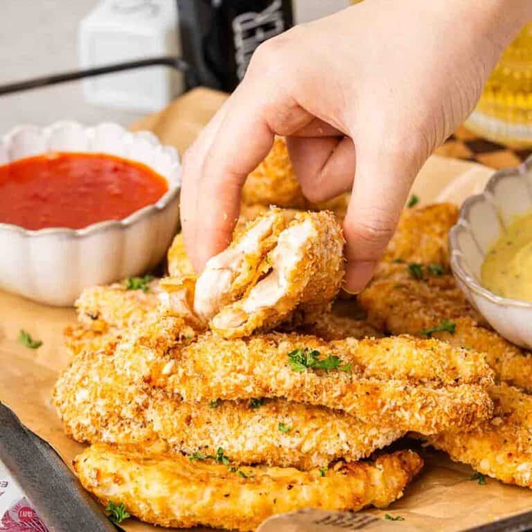 hand holding a chicken tender cut in half over a tray of more chicken and a dipping sauce