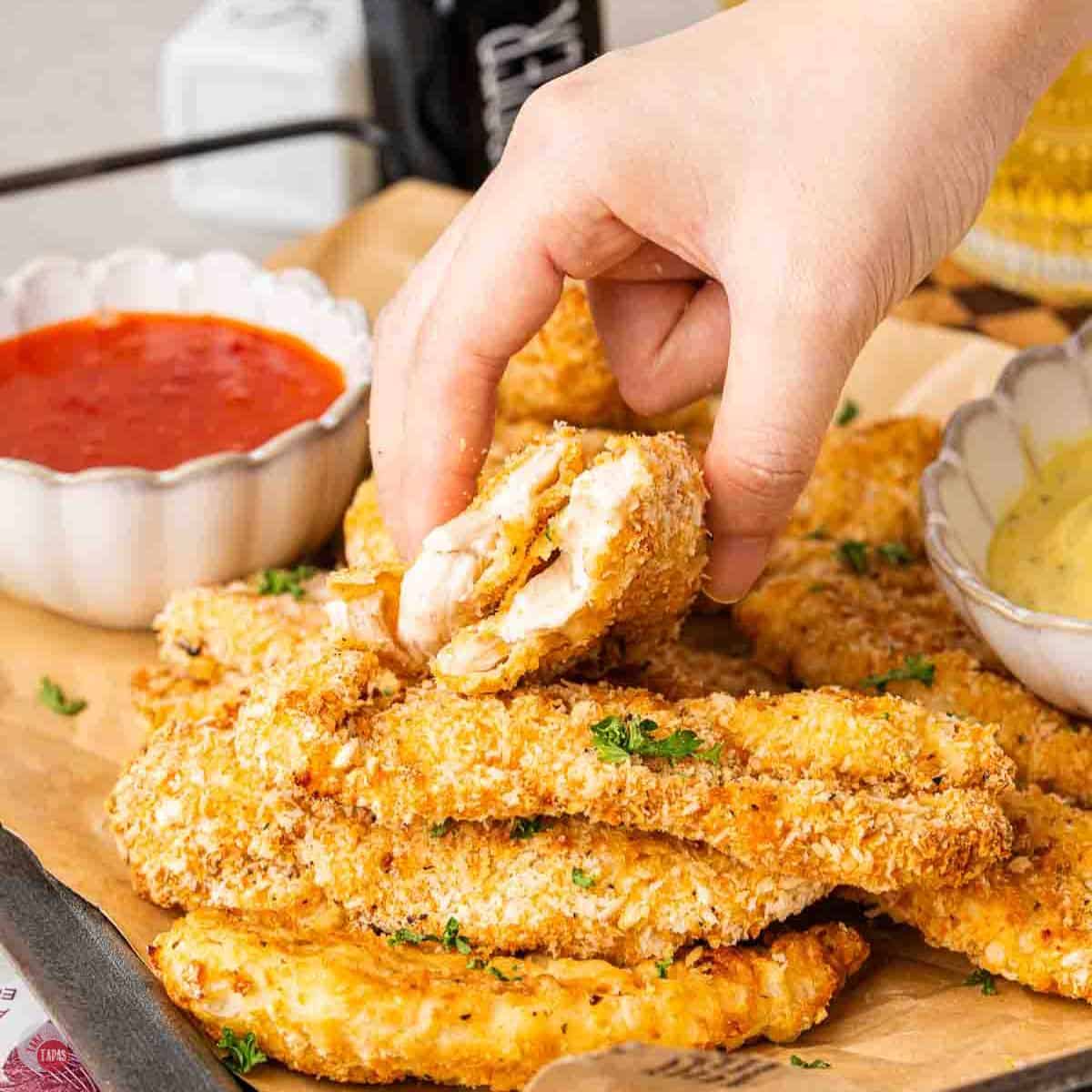hand holding a chicken tender cut in half over a tray of more chicken and a dipping sauce