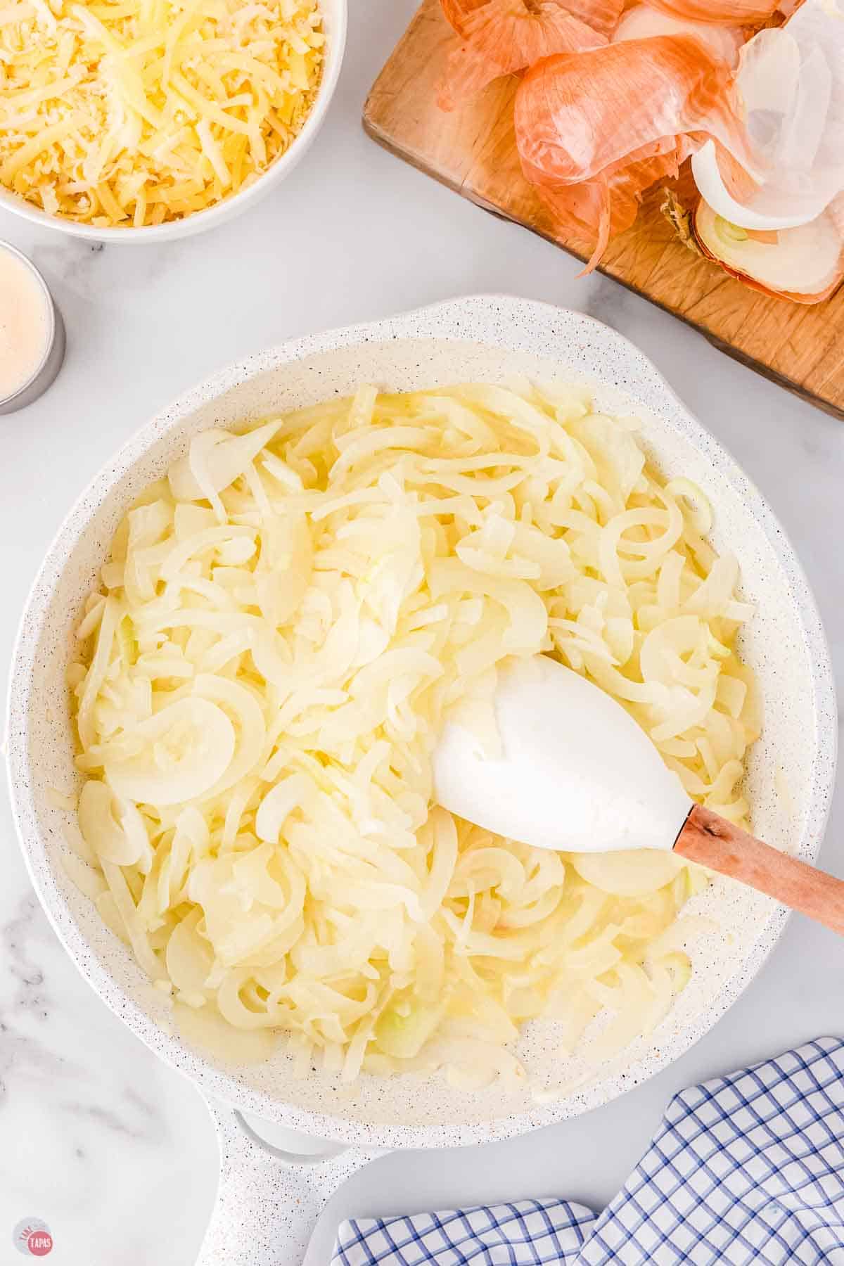 sweet onions cooking in a white skillet and being stirred with a wood spoon