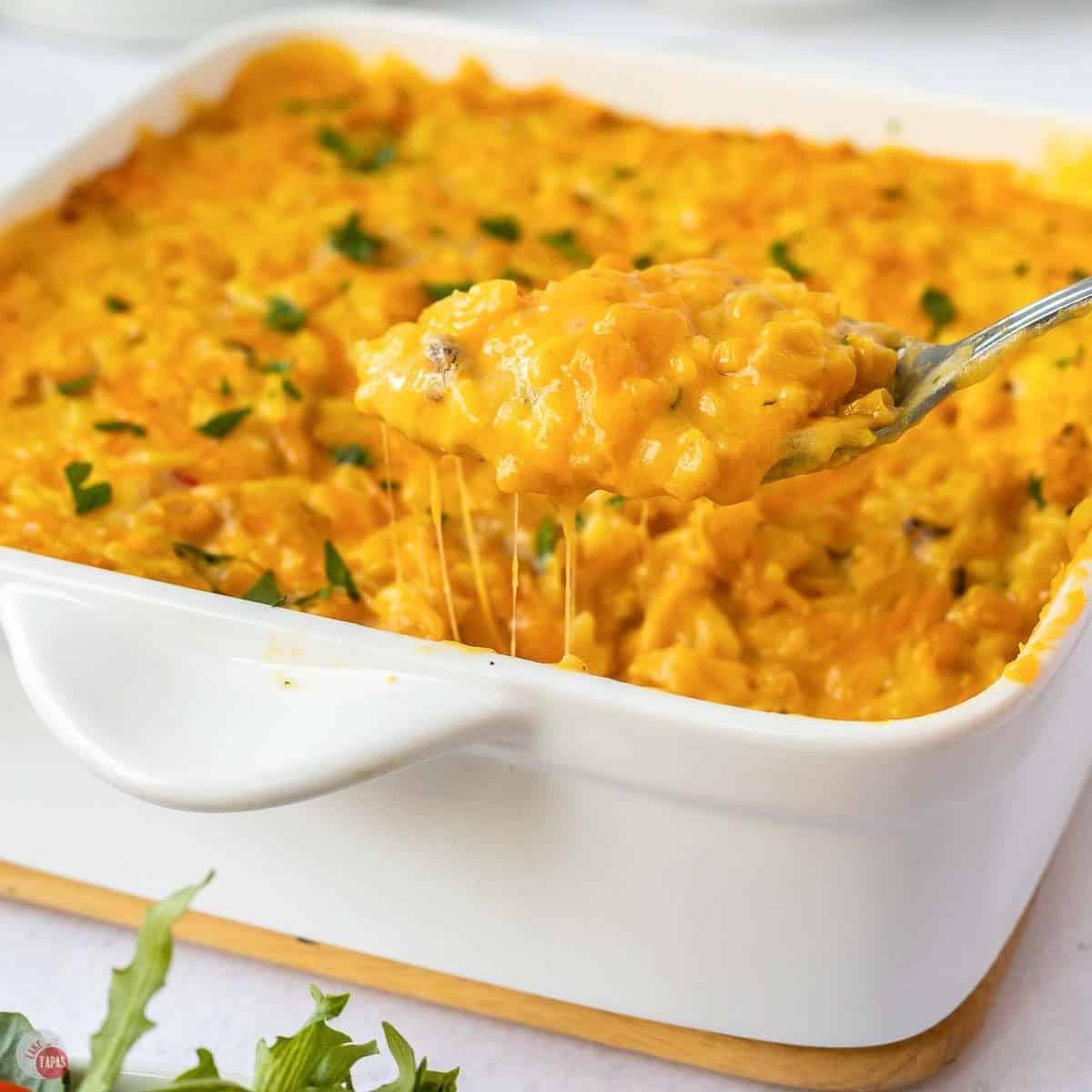 Square dish of baked corn and rice casserole with a serving spoon scooping from one corner