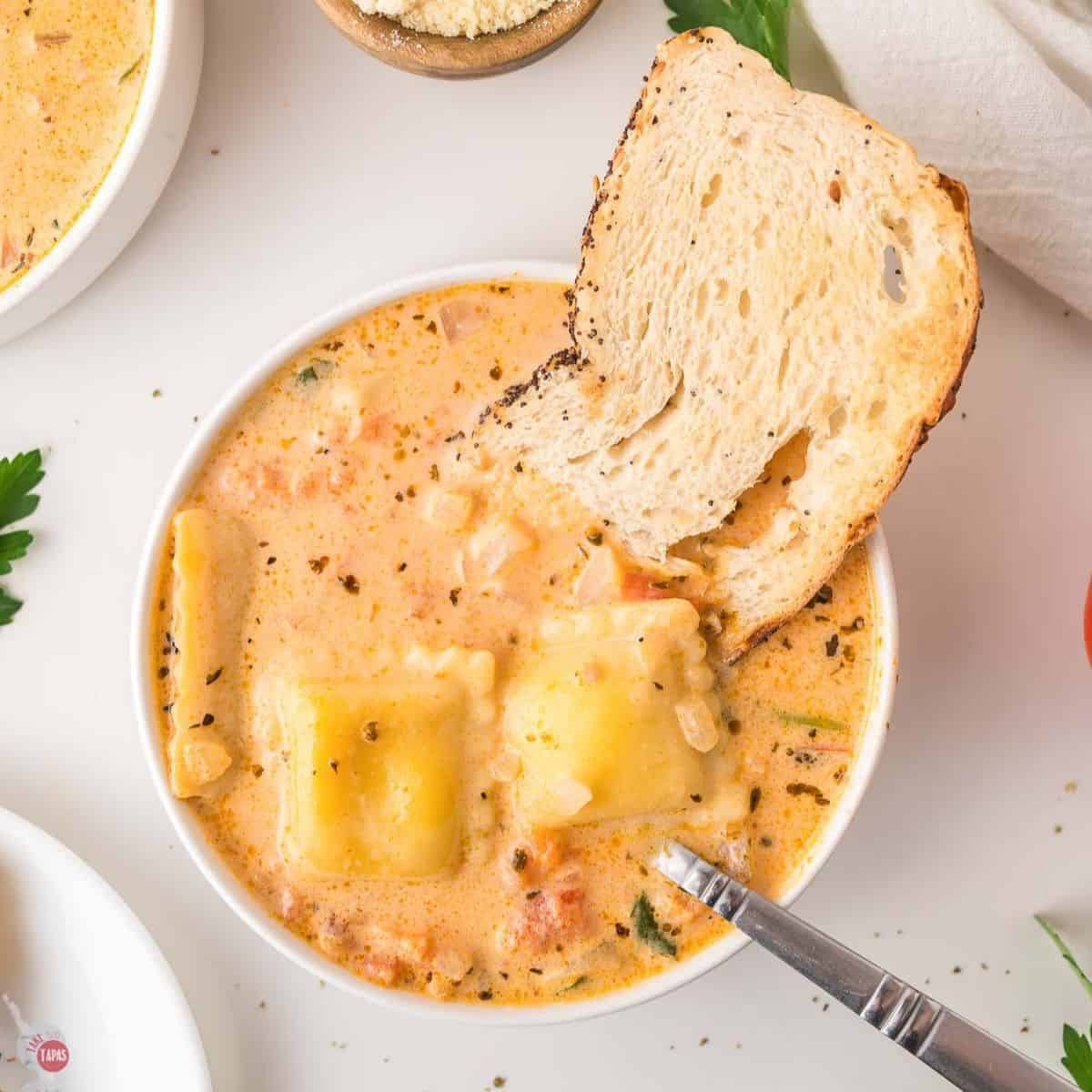 Bowl of creamy ravioli soup with a slice of bread dunked in and a spoon resting in the bowl