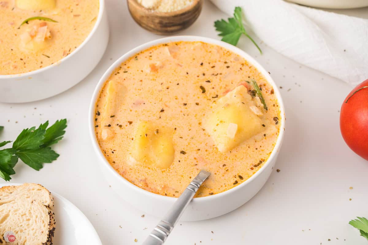 Bowl of creamy tomato ravioli soup with a spoon, a slice of bread and a fresh tomato on the side