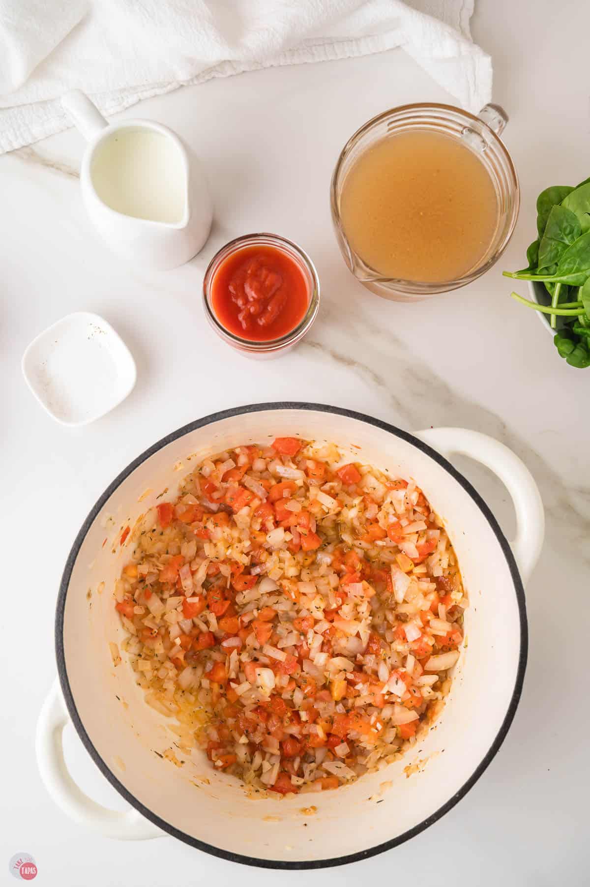 Sauteed onion, garlic and diced tomatoes in a white Dutch oven, the first step of ravioli soup, with broth, cream and tomato sauce nearby